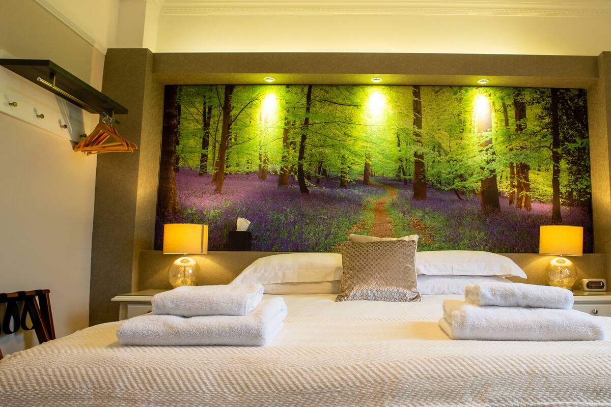 Habitación de hotel con un mural grande de temática forestal en la pared, una cama bien hecha con dos toallas, dos lámparas de noche y una mesita de noche con una lámpara y un reloj.