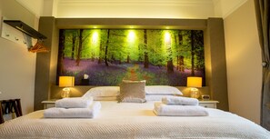 A hotel room with a large forest-themed mural on the wall, a neatly made bed with two towels, two bedside lamps, and a nightstand with a lamp and a clock. - Deanfield Bed and Breakfast (Lydney)