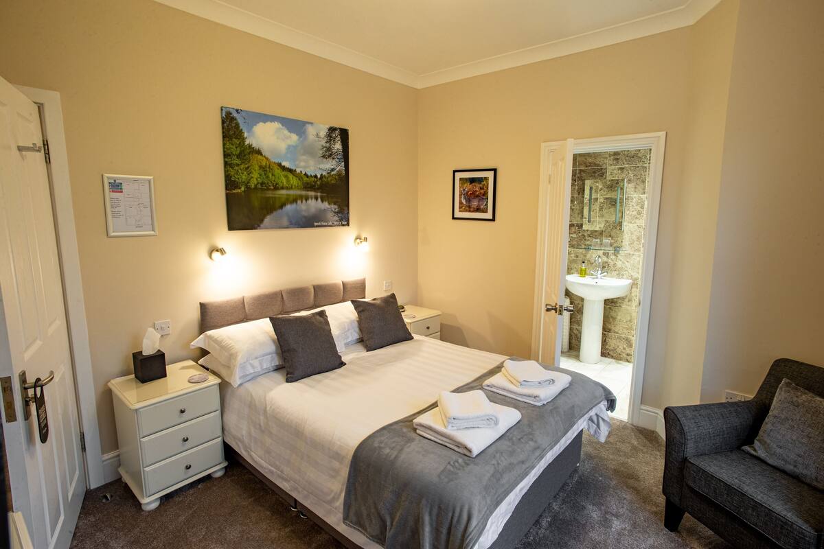 Un dormitorio con cama, mesitas de noche, un sillón, un cuadro, una fotografía enmarco y un baño visible a través de una puerta abierta.