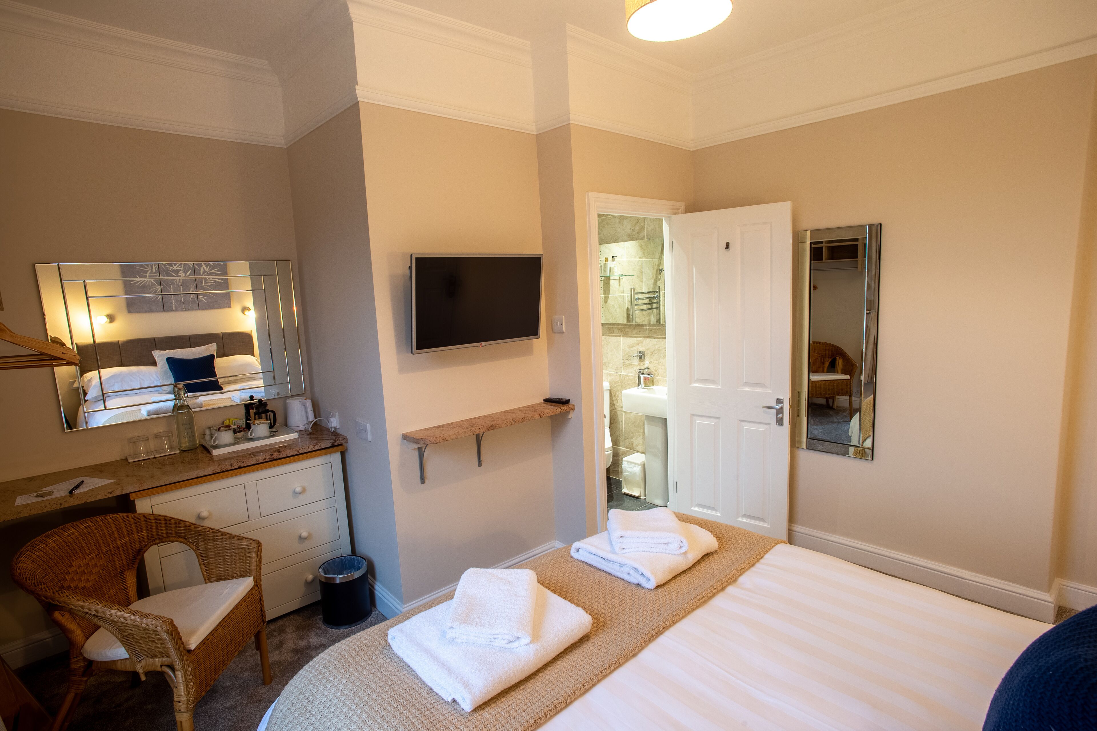 A hotel room with a bed, a television, a bathroom visible through an open door, a wicker chair, and a small shelf with toiletries.