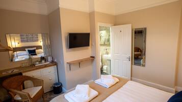 A hotel room with a bed, a television, a bathroom visible through an open door, a wicker chair, and a small shelf with toiletries.