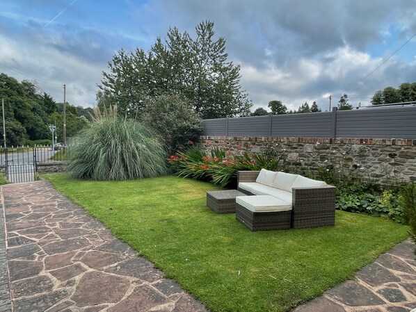A patio area with a stone pathway, a grassy lawn, and a wicker sofa with cushions. - Deanfield Bed and Breakfast (Lydney)