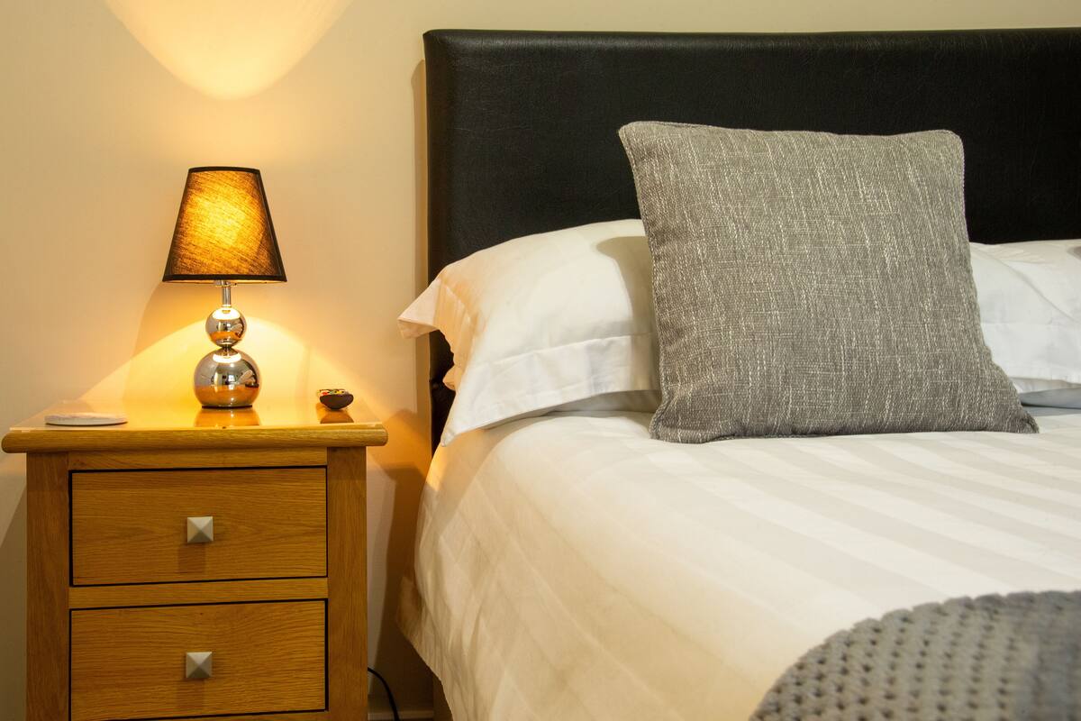 Una cama bien hecha con un almohadón gris, una lámpara de noche y una mesita de noche de madera con cajones.