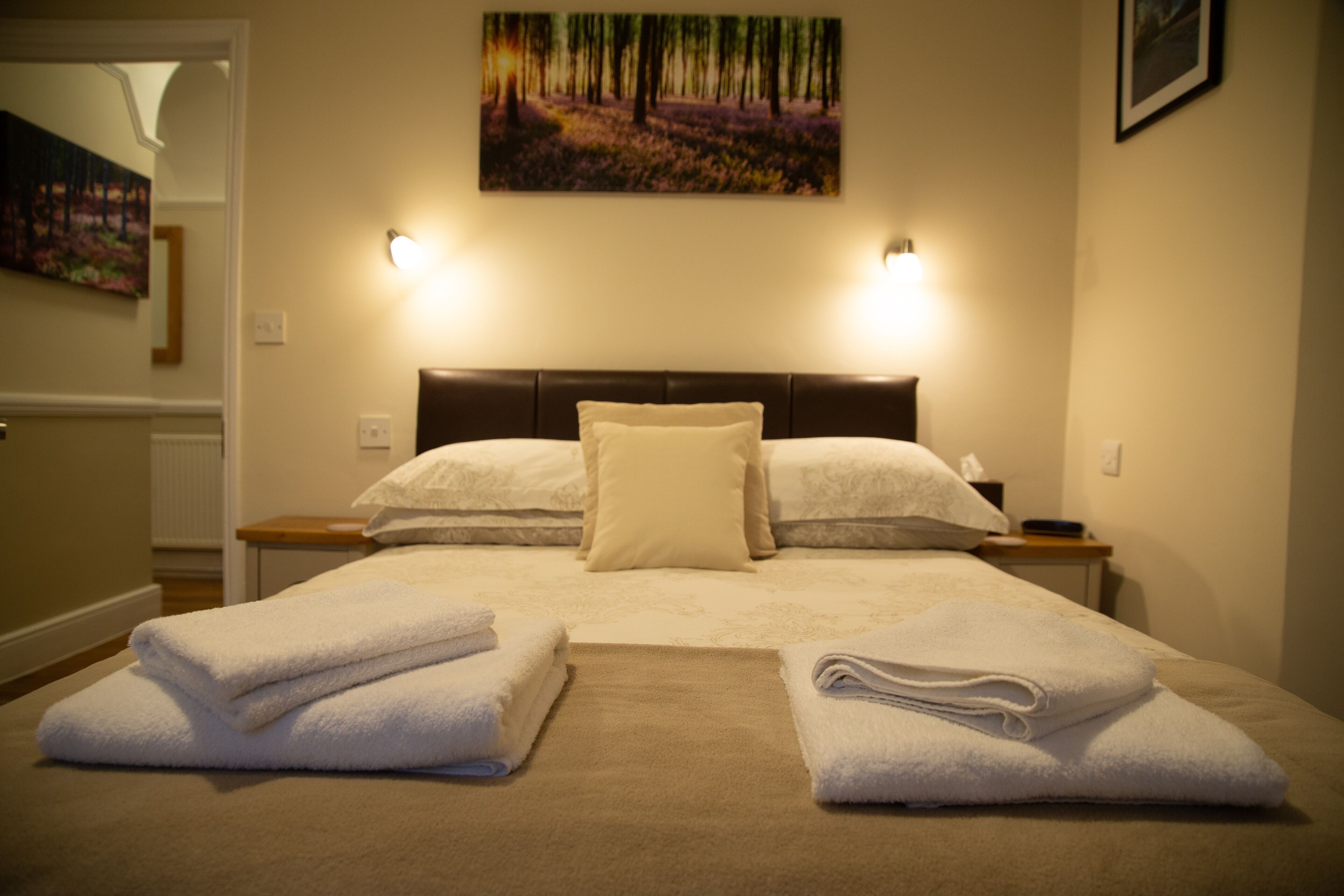 A neatly made bed with white linens and two folded towels on the bed. There is a headboard with a light switch and a small table beside the bed.