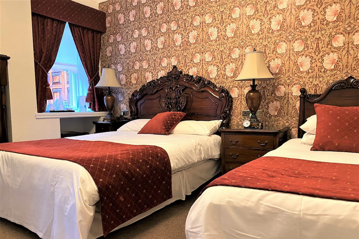 A hotel room with two beds, a nightstand with lamps, a wooden headboard, and a window with curtains.
