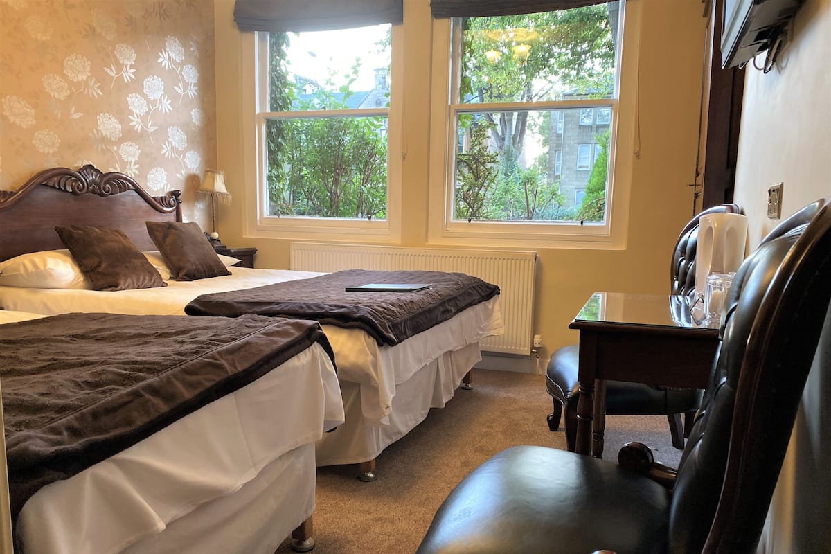 A hotel room with two beds, a desk, and a chair. There are windows with a view of greenery outside.