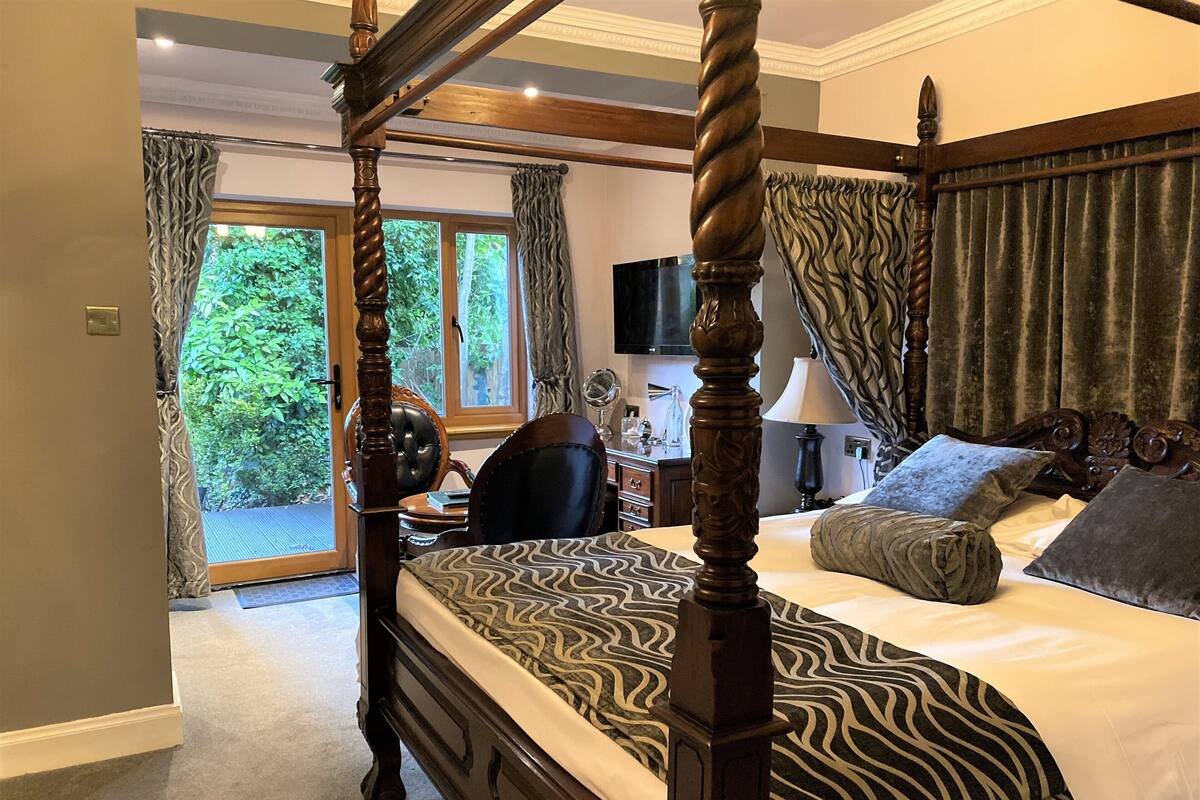 A four-poster bed with a patterned bedspread, a bedside table with a lamp, and a view of greenery through the window.