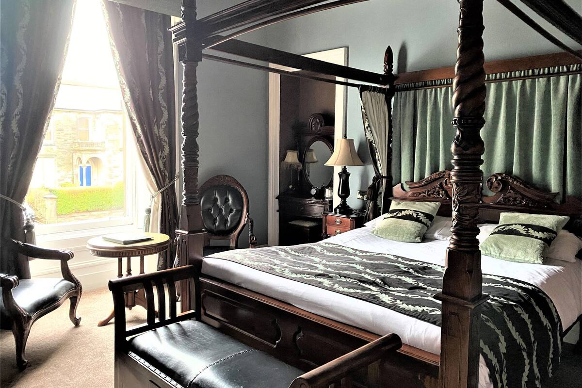 A four-poster bed with a headboard, a bedside table, a chair, and a view of a building through a window.