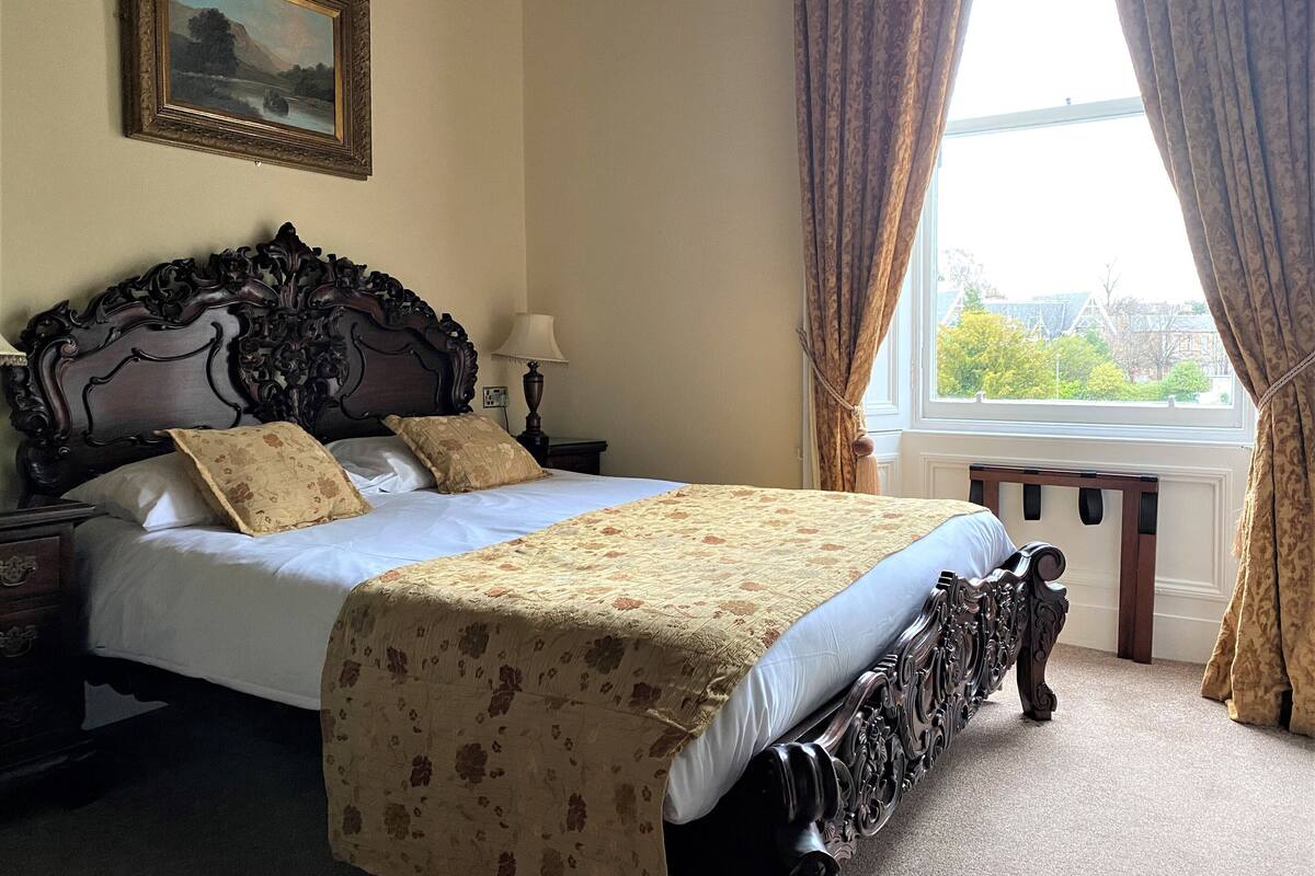 A bedroom with a large bed, ornate headboard, a nightstand with a lamp, and a window with curtains.