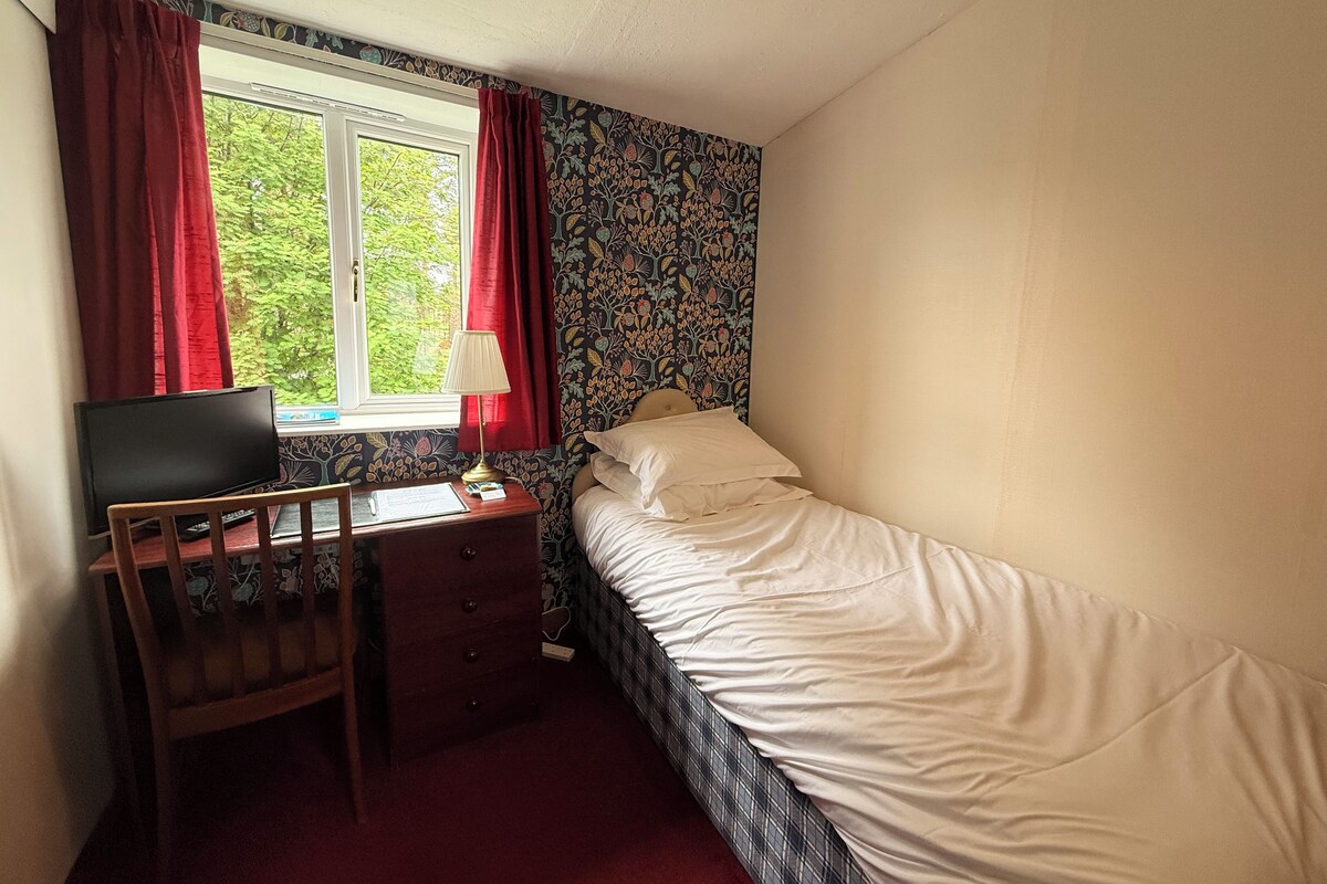 Une chambre avec un lit simple, un bureau avec une chaise, une télévision et une fenêtre avec des rideaux rouges.