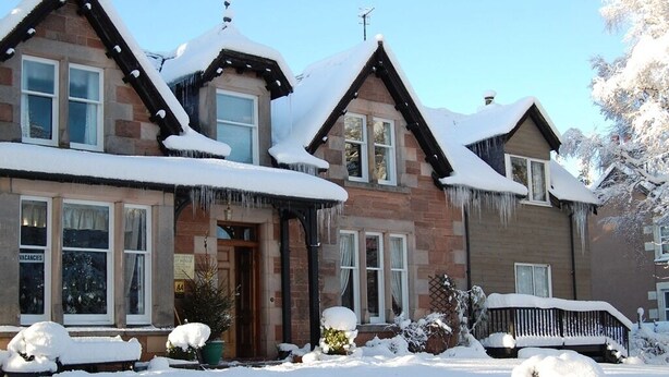 Une maison à deux étages, dont le toit est recouvert de neige, et sur laquelle on peut voir une pancarte indiquant « Chambres disponibles » sur la porte d’entrée.