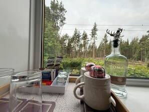 Une table avec une bouteille de whisky écossais, une tasse et quelques barres de chocolat, placée près d’une fenêtre donnant sur une forêt.