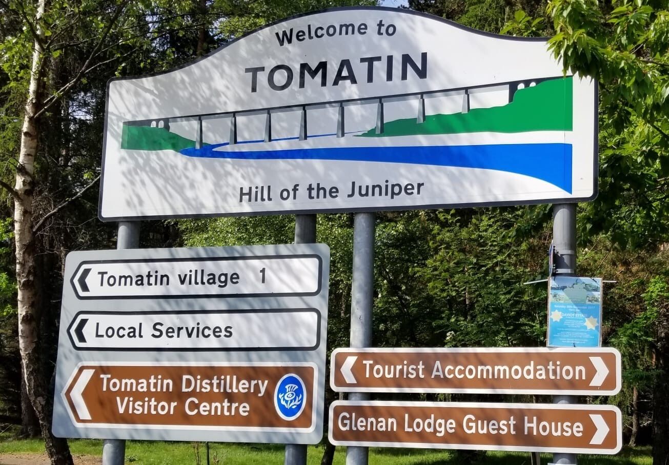A signpost with directions to Tomatin village, local services, Tomatin Distillery Visitor Centre, and tourist accommodations including Glenan Lodge Guest House.