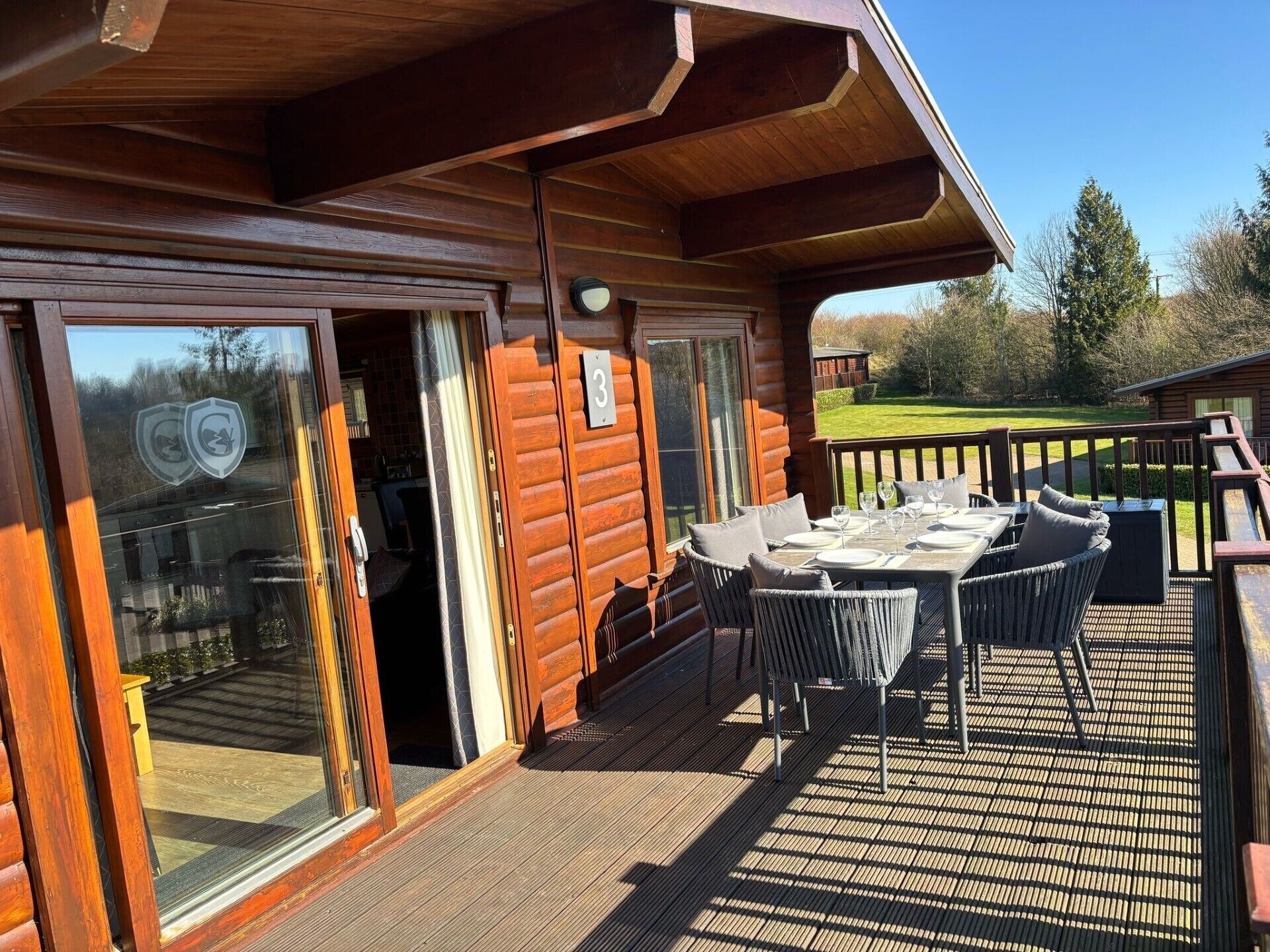 Une terrasse en bois avec une table et des chaises, une porte vitrée donnant sur une pièce et une enseigne portant le chiffre « 3 ».