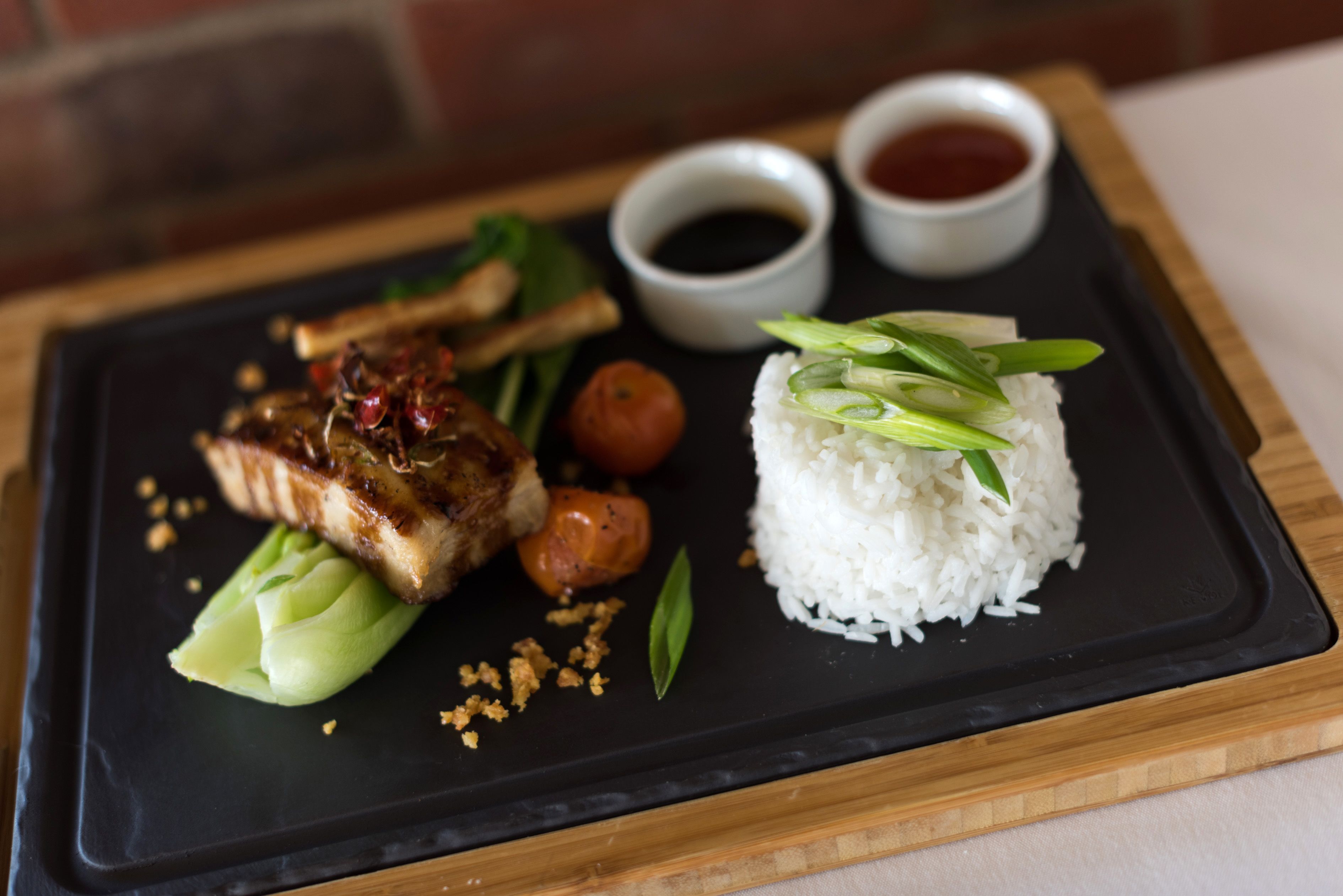 A black slate plate with white rice, grilled meat, green onions, and two small bowls of sauce.