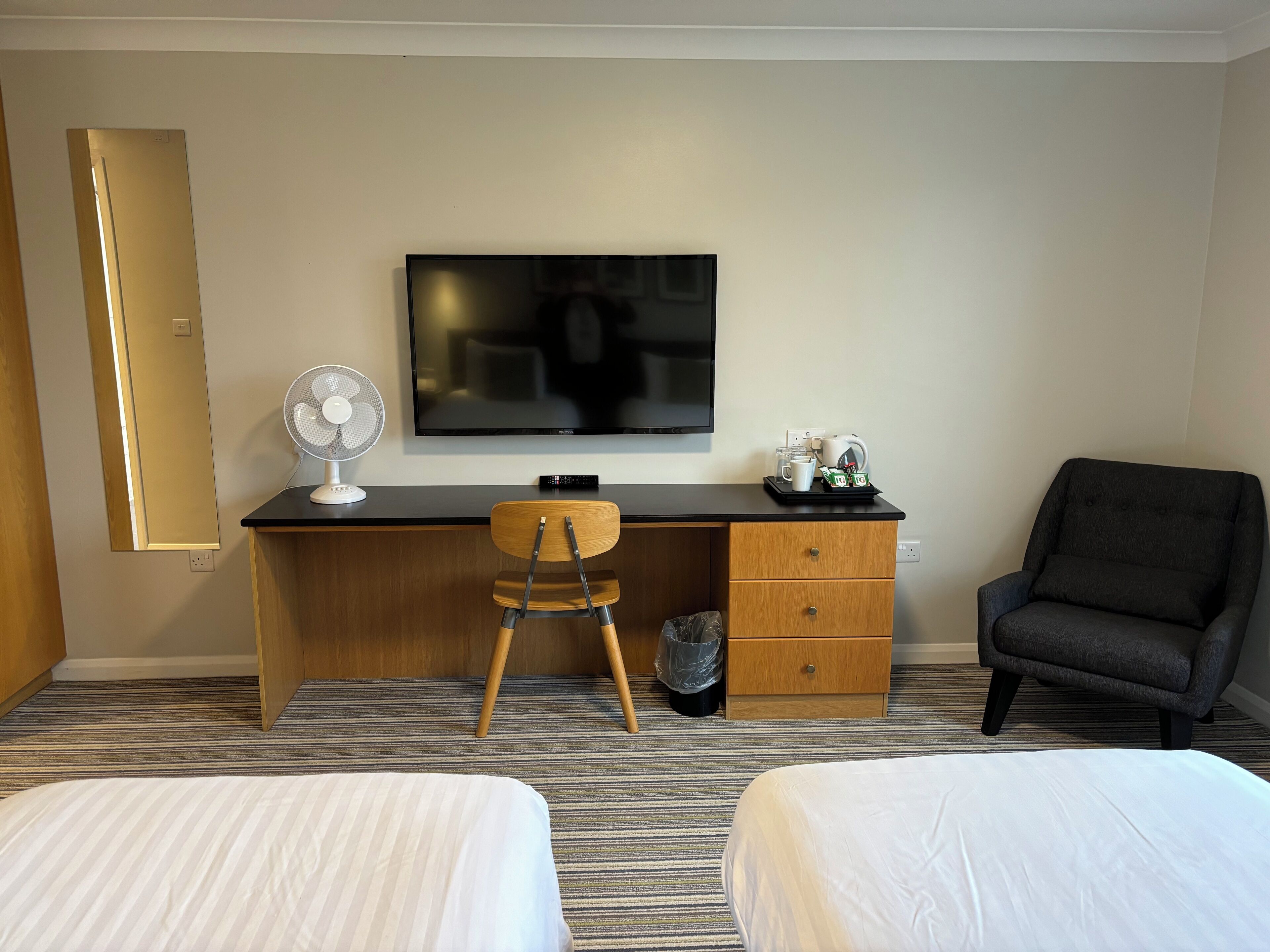 Ein Hotelzimmer mit Bett, Schreibtisch mit Stuhl, Fernseher, Ventilator und Sessel.