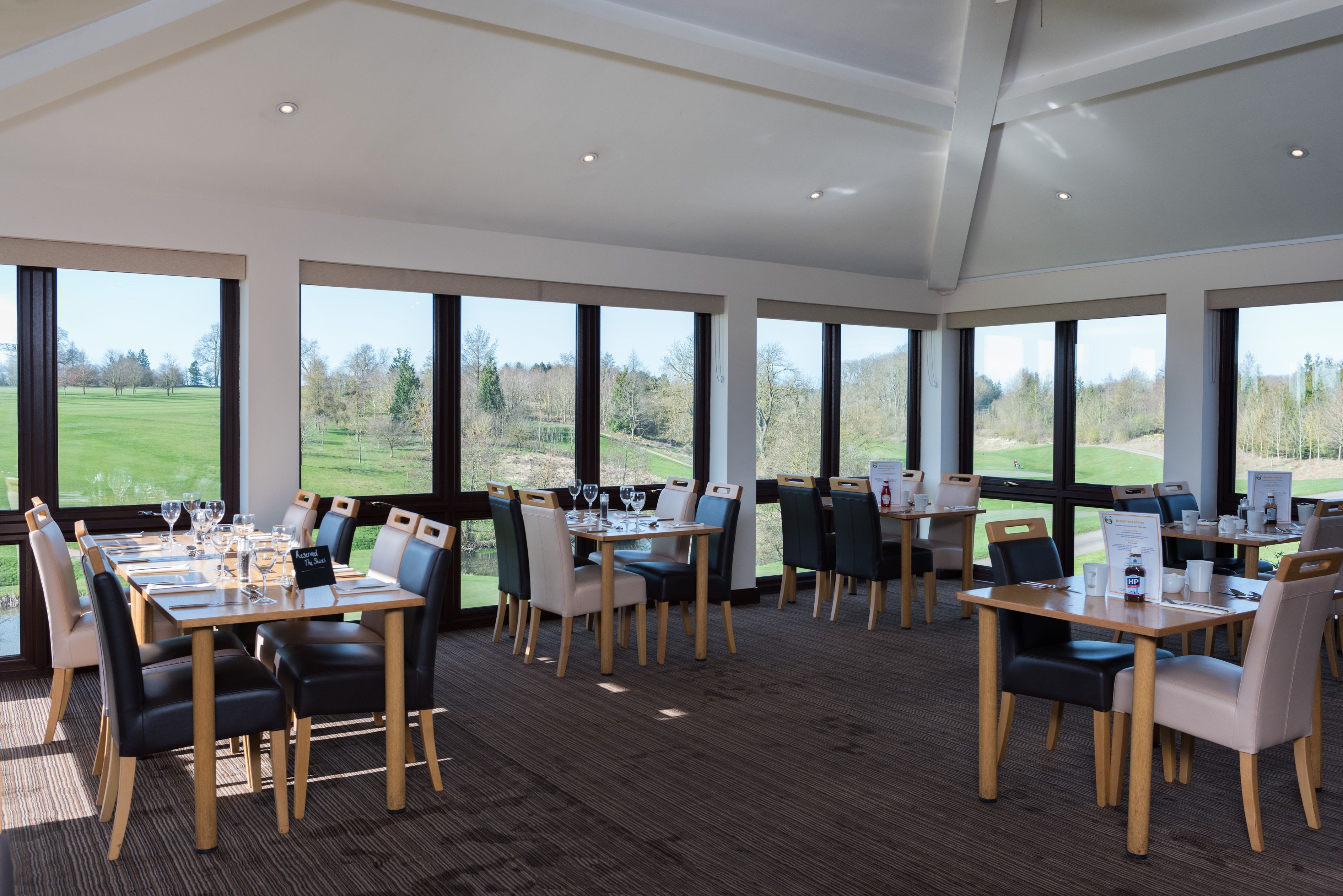 A dining area with tables set for a meal, large windows offering a view of a golf course, and a high ceiling with recessed lighting.