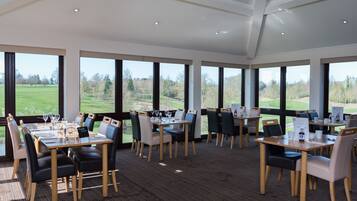 A dining area with tables set for a meal, large windows offering a view of a golf course, and a high ceiling with recessed lighting.