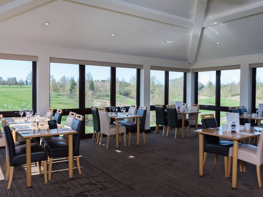 A dining area with tables set for a meal, large windows offering a view of a golf course, and a high ceiling with recessed lighting.
