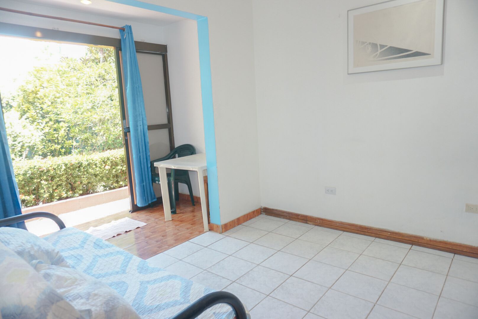 A bedroom with a bed, a small table, and a chair. There is a window with a view of greenery outside.