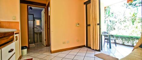 A room with a tiled floor, a bed, a small table, and a chair. There is a door leading to a bathroom and a window with curtains.