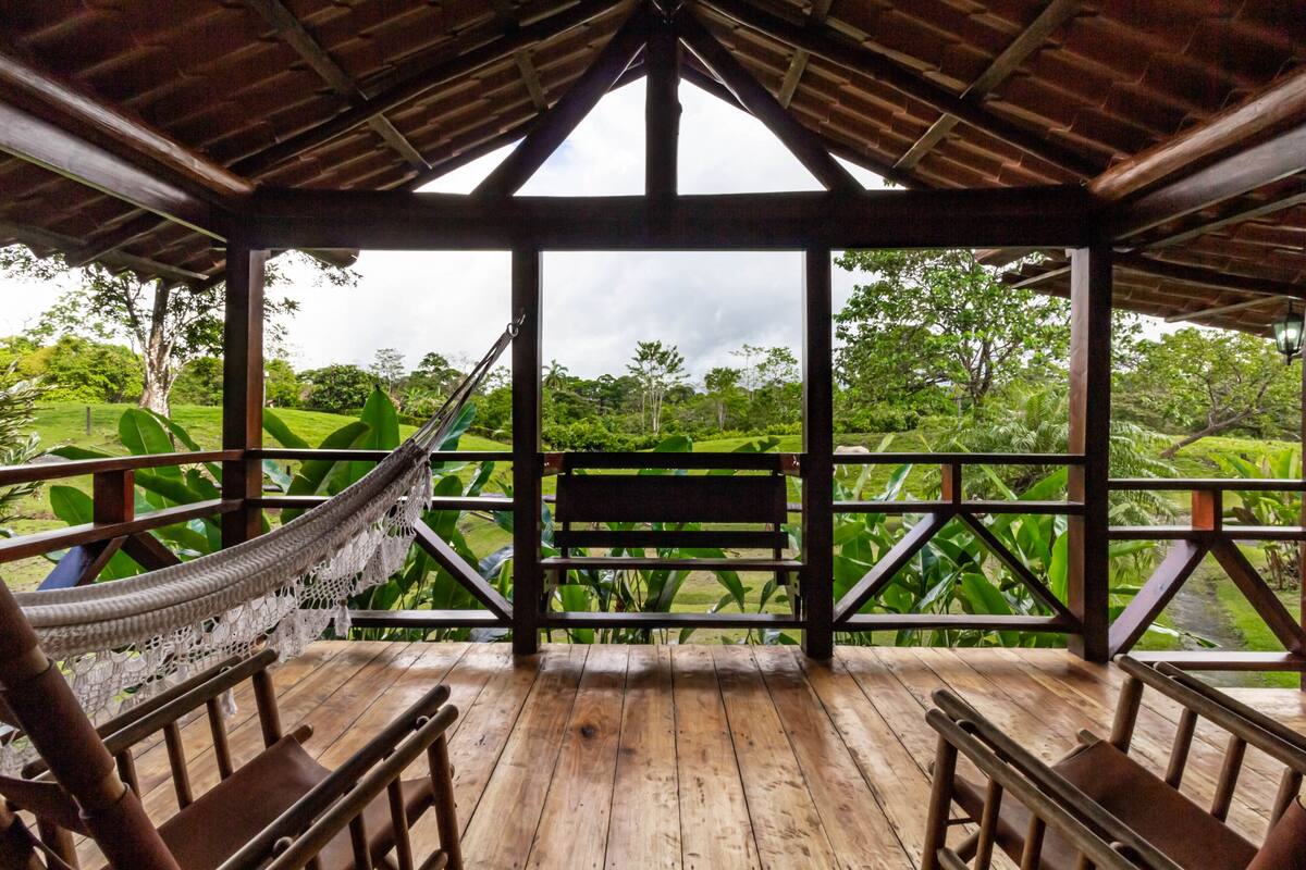 A wooden deck with a hammock and benches, overlooking a lush green landscape.
