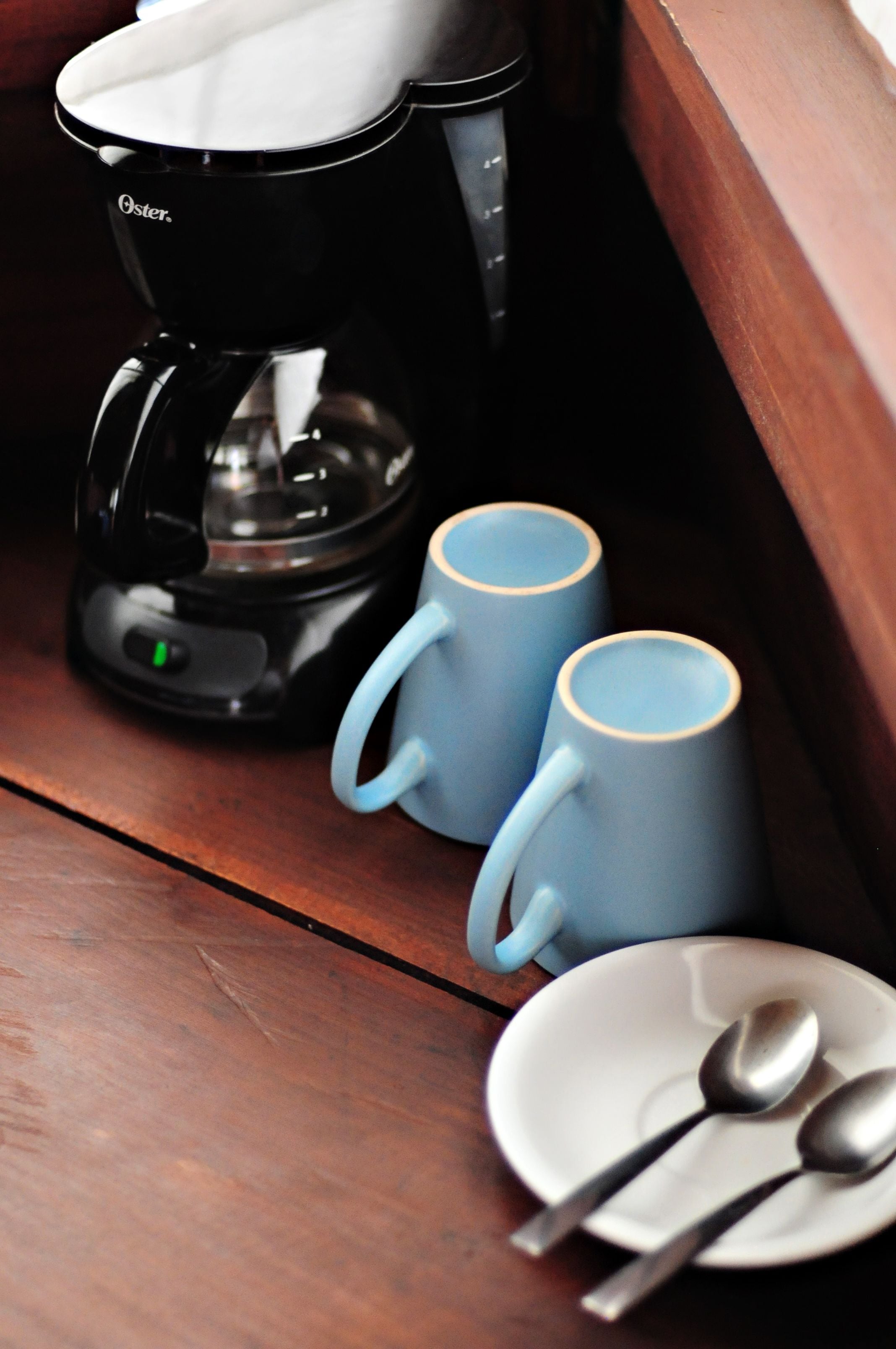 Eine Kaffeemaschine, zwei Tassen und ein Teller mit Löffeln auf einer Holztischplatte.