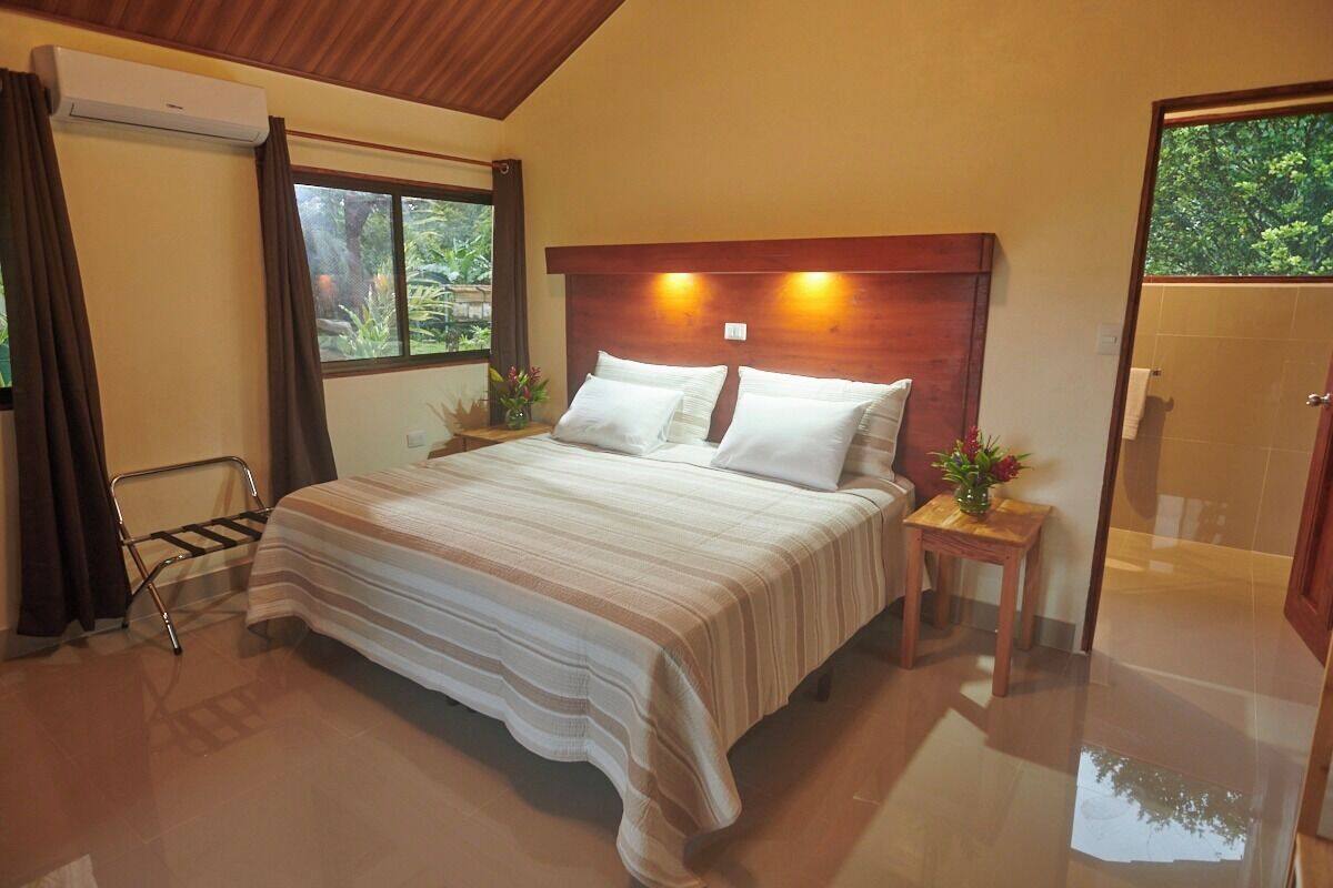 A hotel room with a large bed, a wooden headboard, a window with curtains, a small table with a vase, and a shower area.