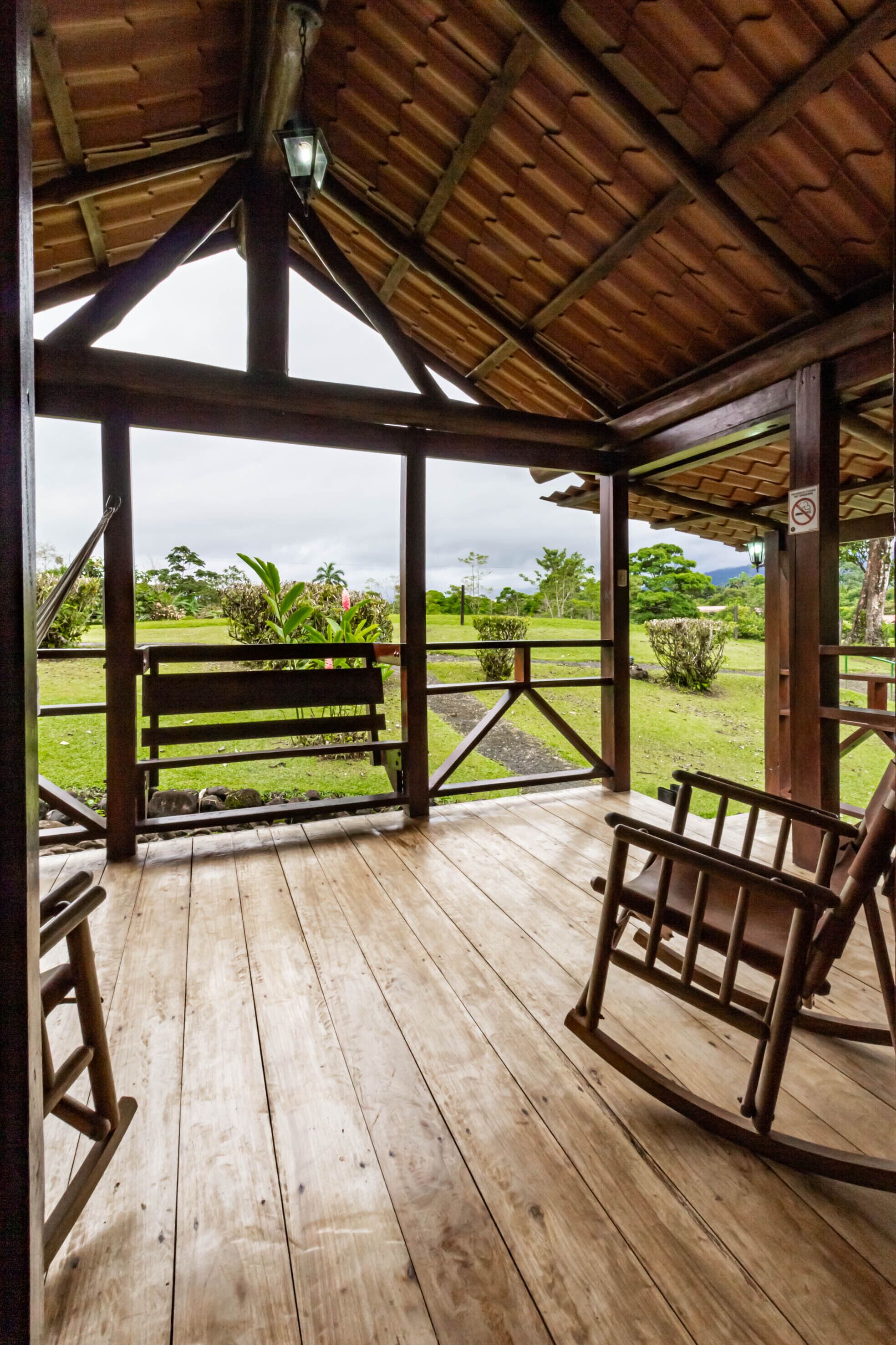 A wooden deck with a bench and a rocking chair, overlooking a grassy area with plants and trees.