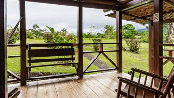 Una terraza de madera con un banco y una mecedora, con vistas a un área verde con plantas y árboles.