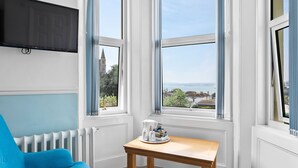 Una habitación con un sillón azul, una mesita de madera con una cafetera y tazas, y un televisor montado en la pared.