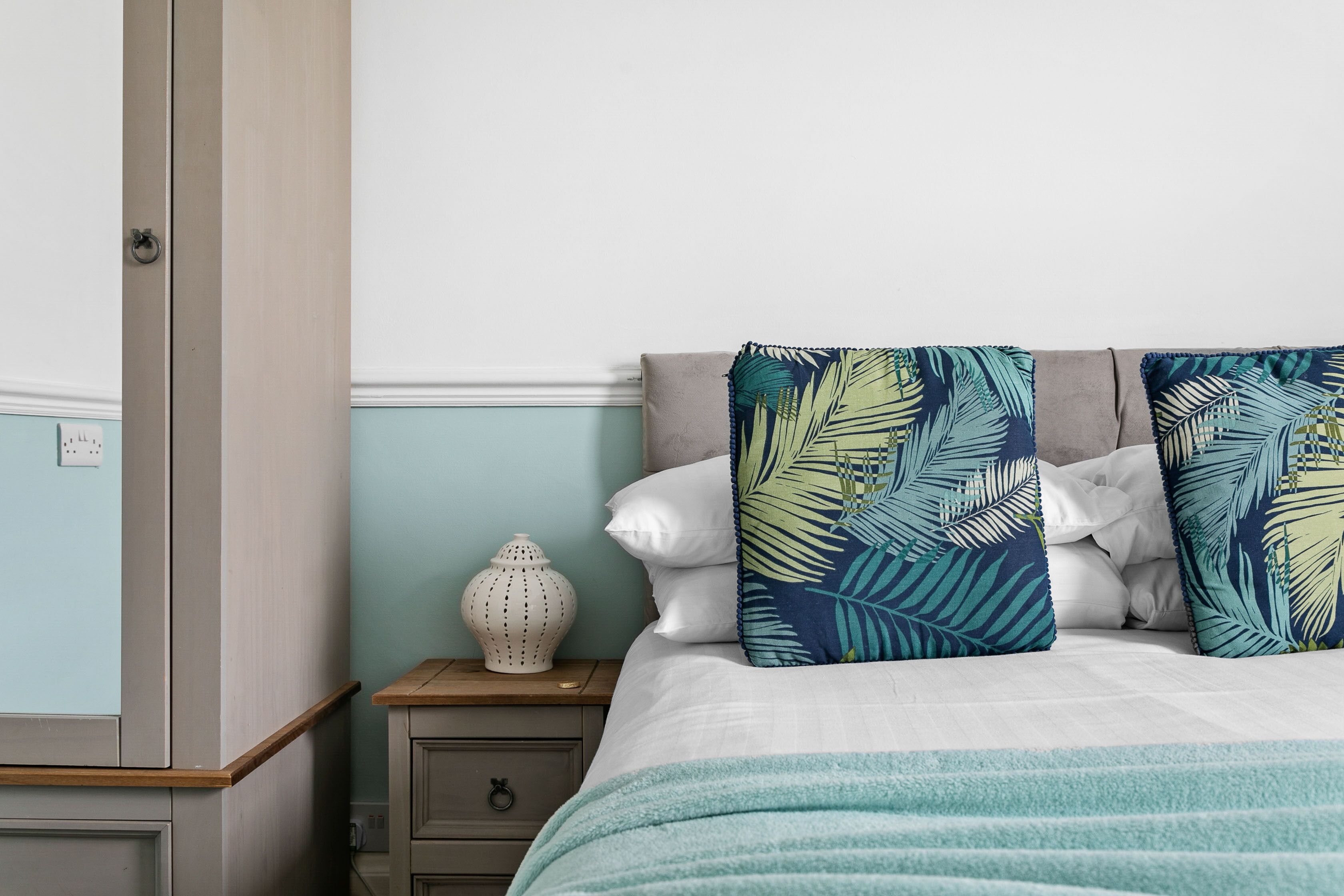 Una cama bien tendida con una manta azul y blanca a rayas, dos almohadas con motivos de hojas tropicales y un jarrón blanco sobre una mesita de noche.
