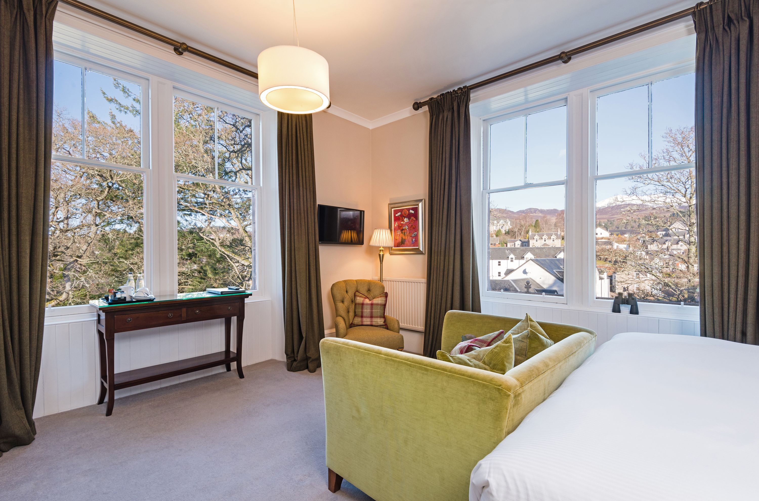 A hotel room with a bed, a green armchair, a wooden desk, and a view of a snowy landscape through large windows.