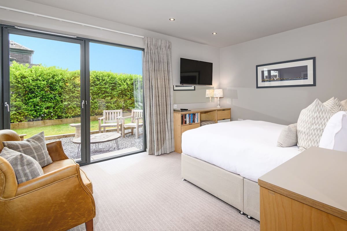 A modern hotel room with a large bed, a desk, a TV, and a sliding glass door leading to an outdoor seating area.
