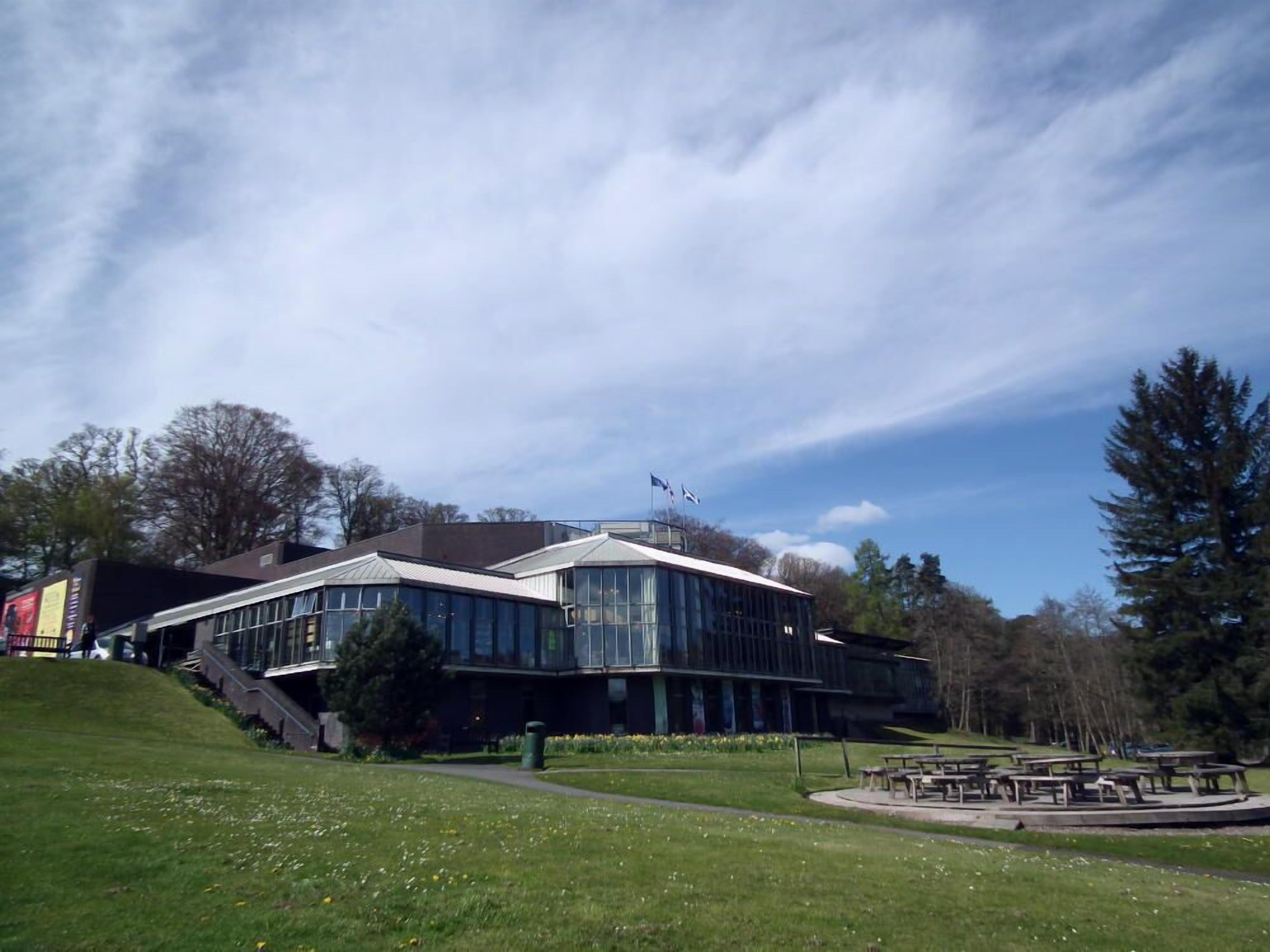 Een modern gebouw met een gebogen glazen gevel, omgeven door een gazon met picknicktafels.