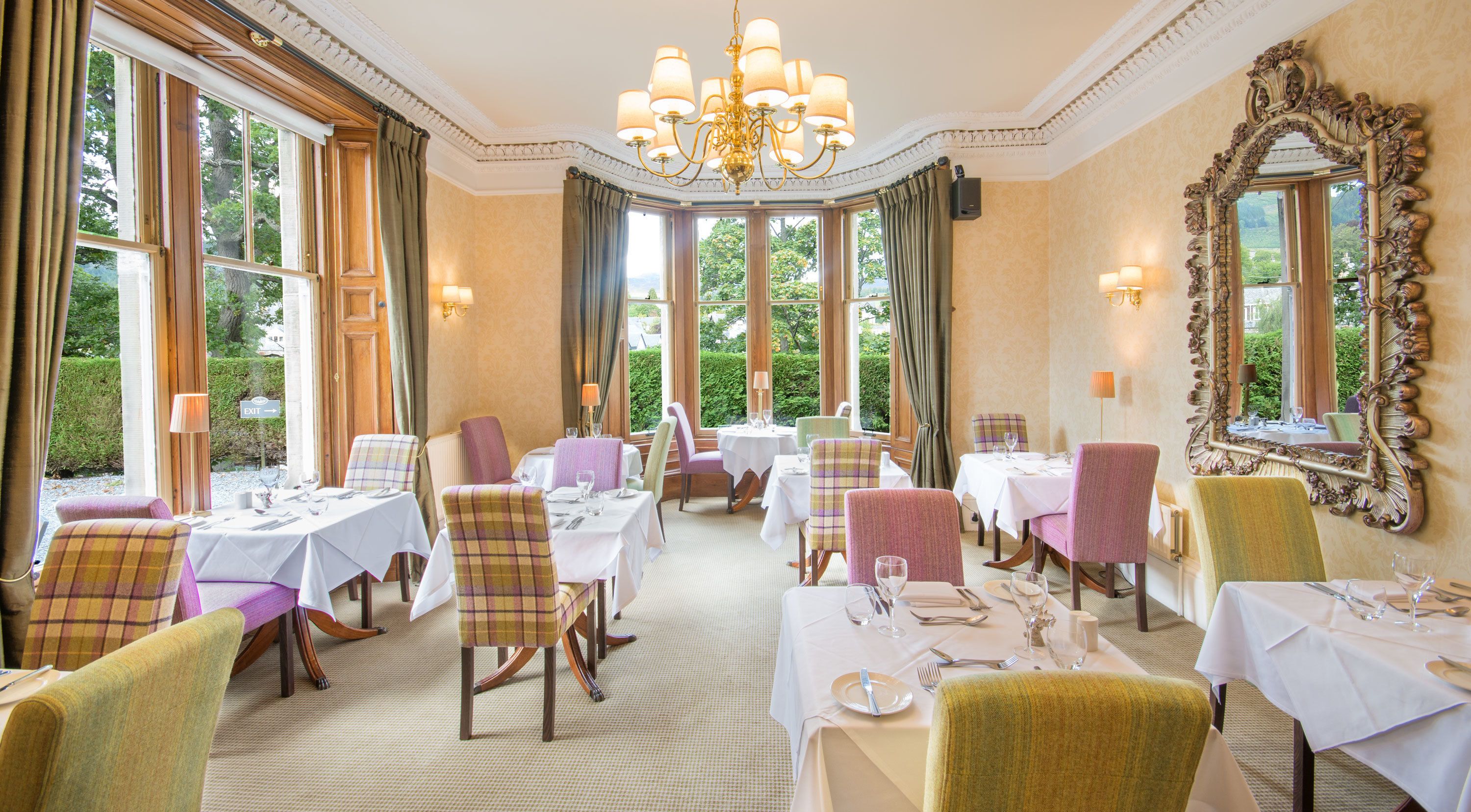 A formal dining room with multiple tables and chairs, large windows with curtains, and a chandelier.