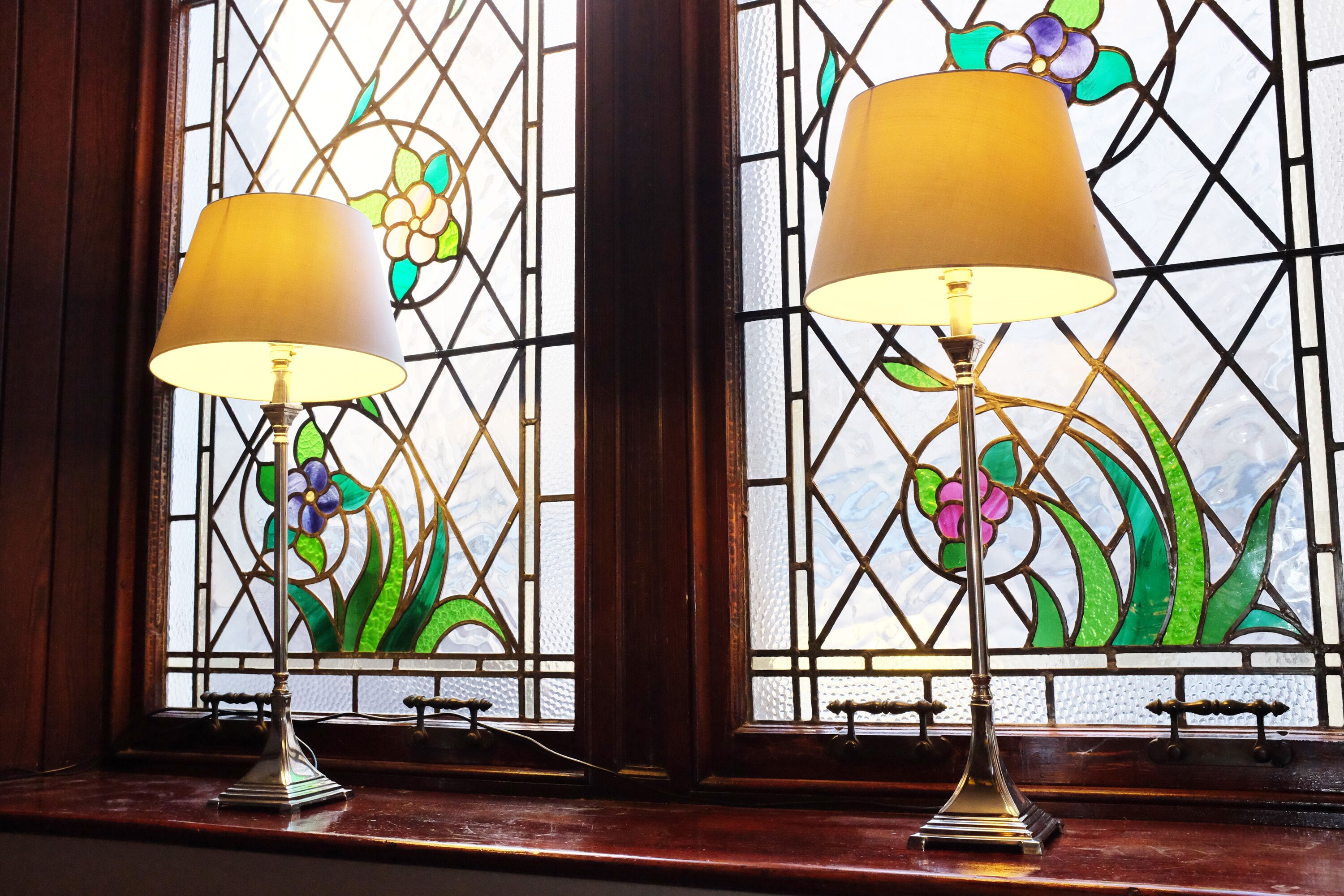 Two floor lamps on a windowsill with stained glass behind them.