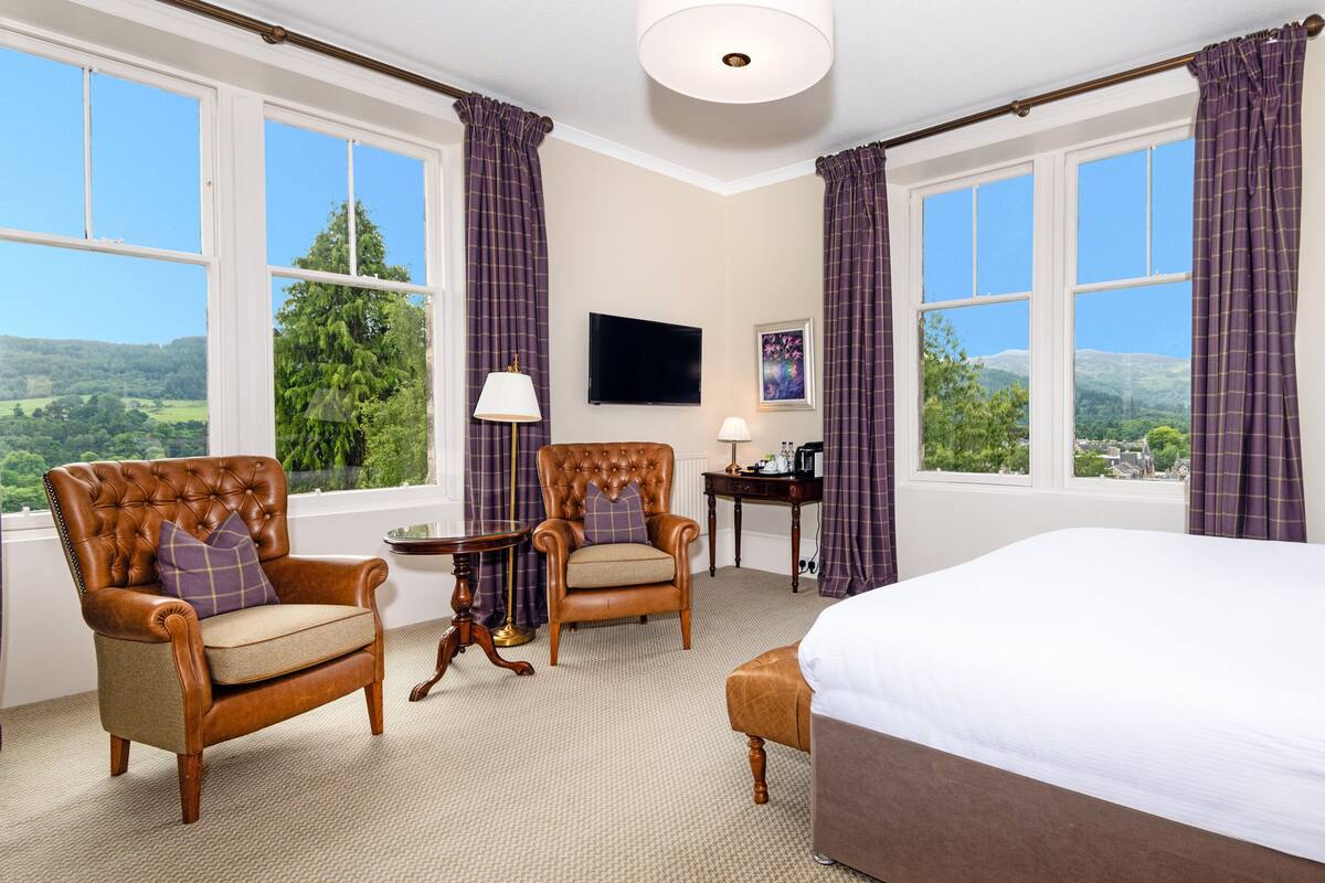 A hotel room with a large bed, two armchairs, a small table, and a desk. There are large windows with curtains, a flat-screen TV, and a painting on the wall.