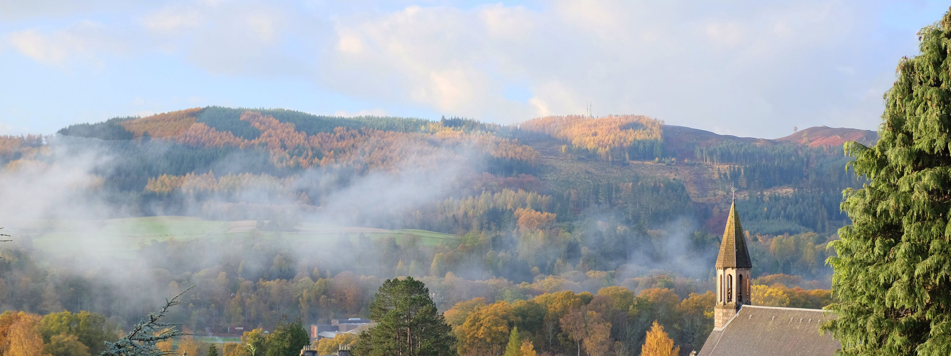 Une église dotée d’un clocher, entourée d’un feuillage automnal et de collines brumeuses.