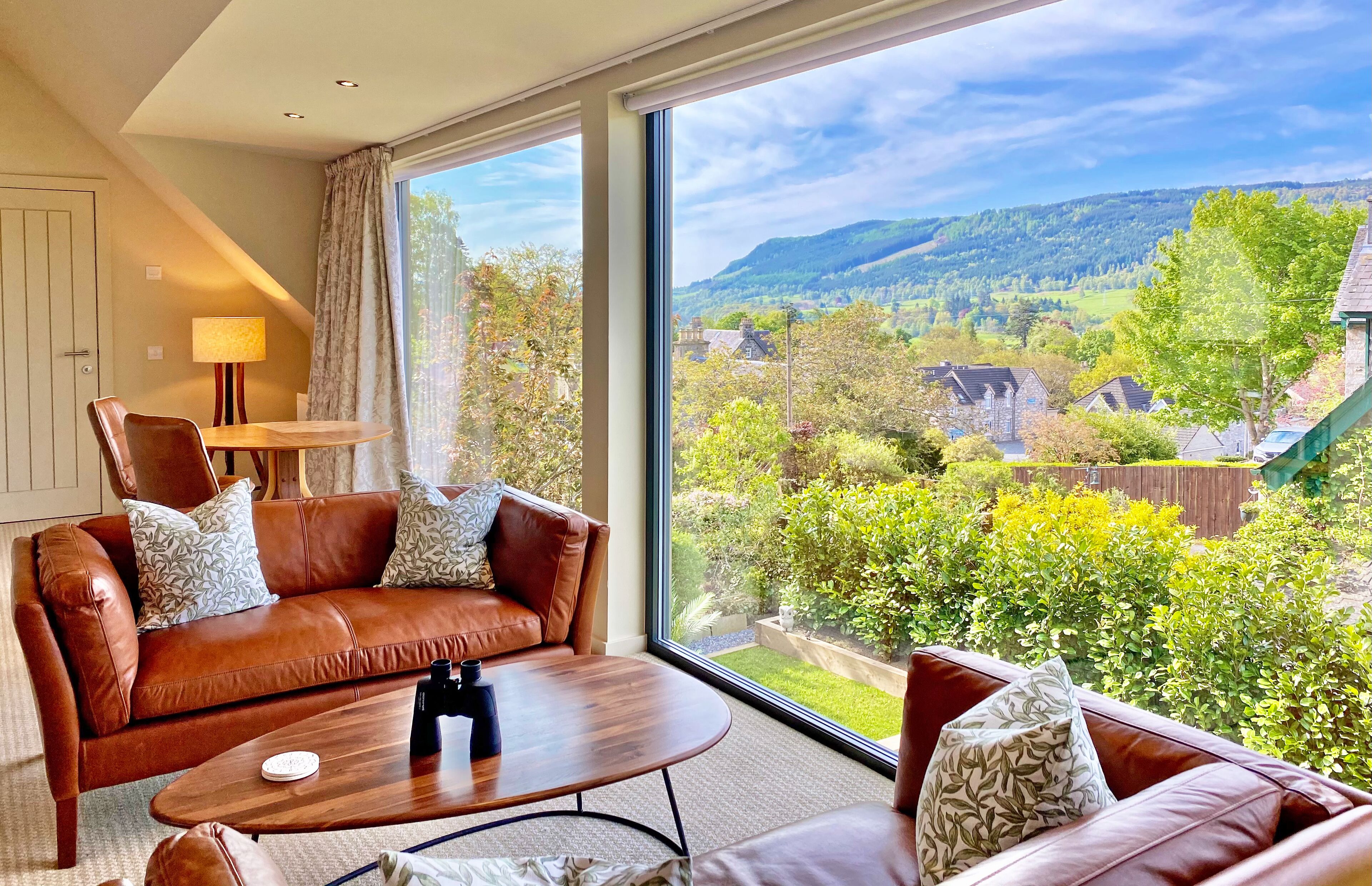 A modern living room with a leather sofa, a wooden coffee table, and a large window offering a scenic view of a green landscape and hills.