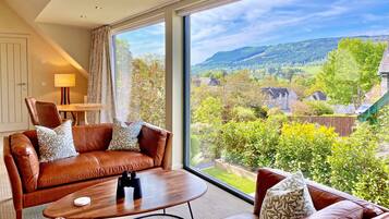 A modern living room with a leather sofa, a wooden coffee table, and a large window offering a scenic view of a green landscape and hills.