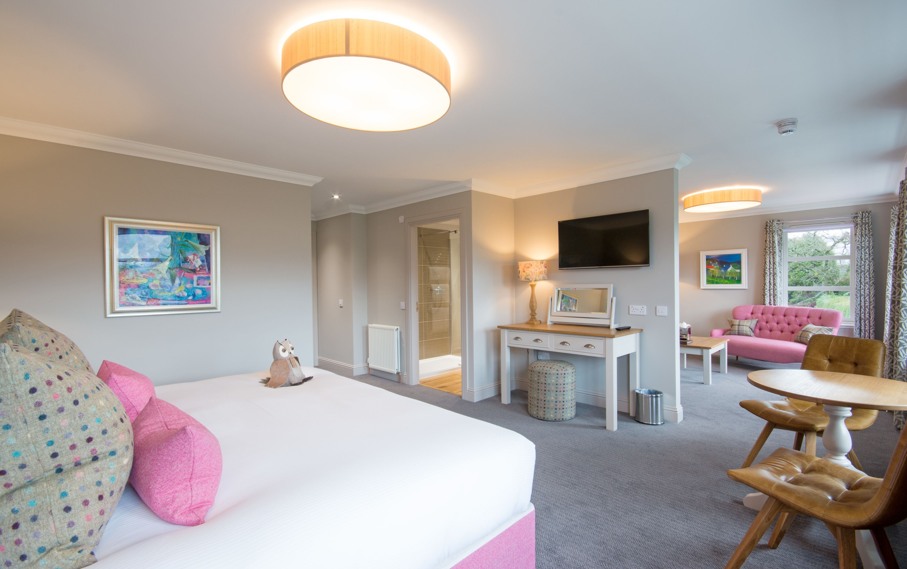 Quarto de hotel com uma cama grande, uma escrivaninha com televisão, um sofá rosa e um espaço de jantar com cadeiras de madeira.