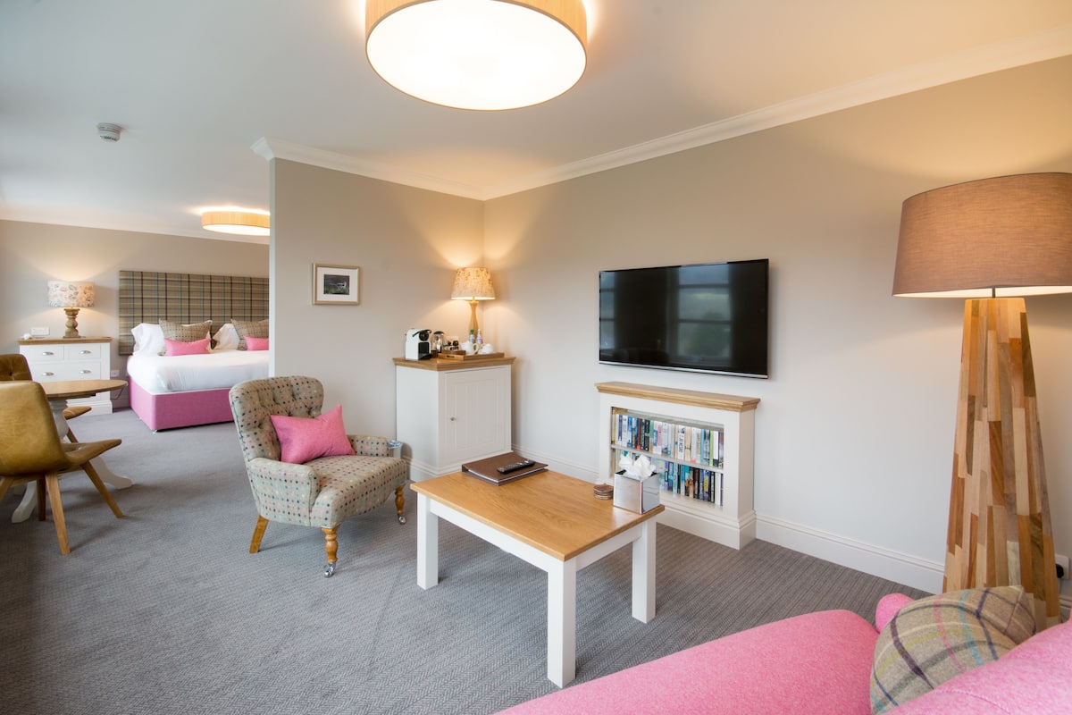 A modern hotel room with a bed, a seating area with a table and chairs, a television, and a bookshelf.