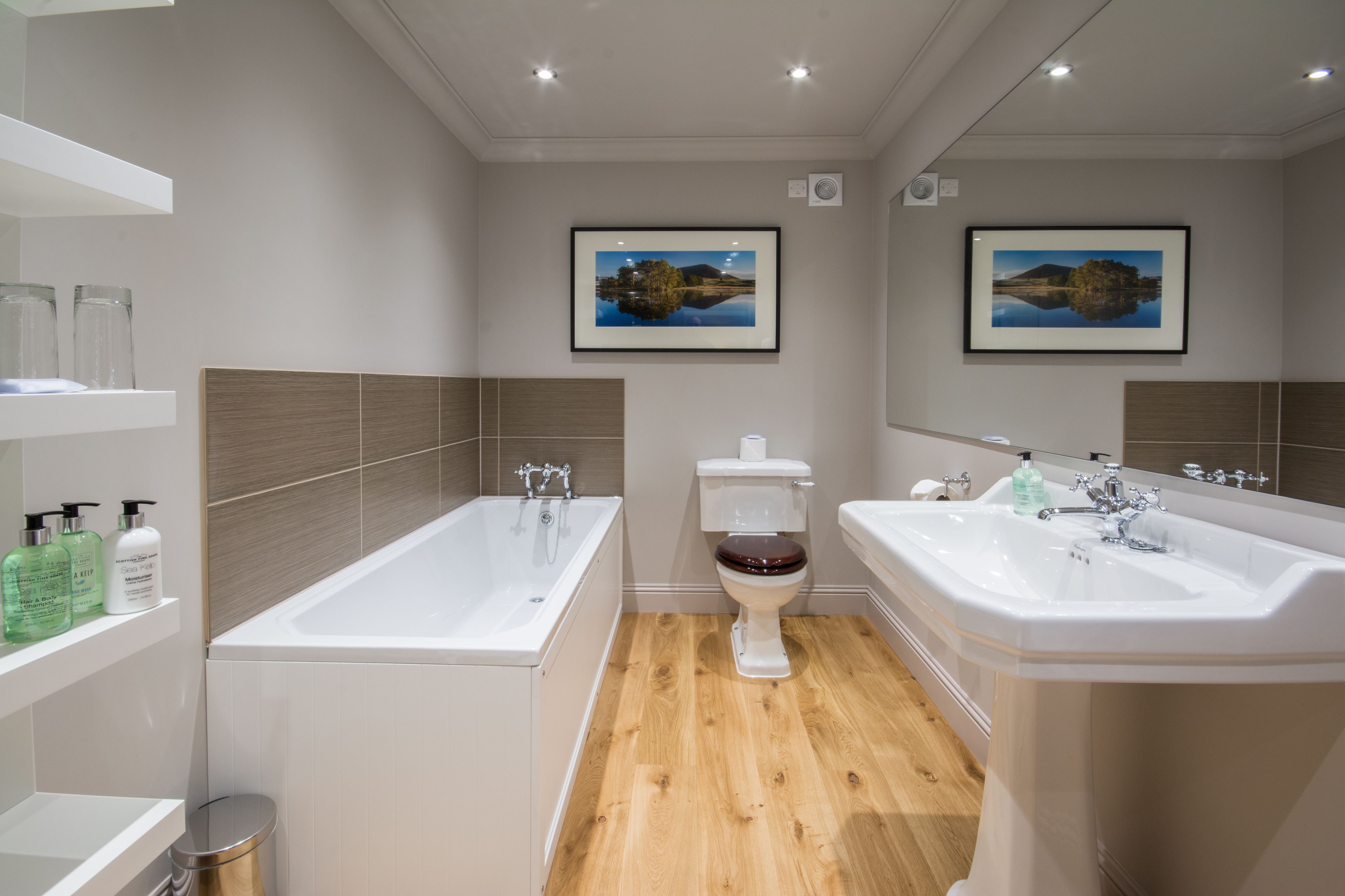 A modern bathroom with a bathtub, toilet, and sink. There are framed pictures on the wall, a shelf with toiletries, and a trash can.