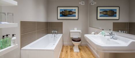 A modern bathroom with a bathtub, toilet, and sink. There are framed pictures on the wall, a shelf with toiletries, and a trash can.