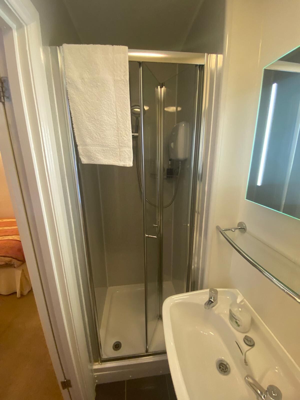 Una ducha con cerramiento de vidrio, una toalla blanca colgada en la puerta, una puerta de vidrio para la ducha y un lavamanos blanco con un espejo arriba.