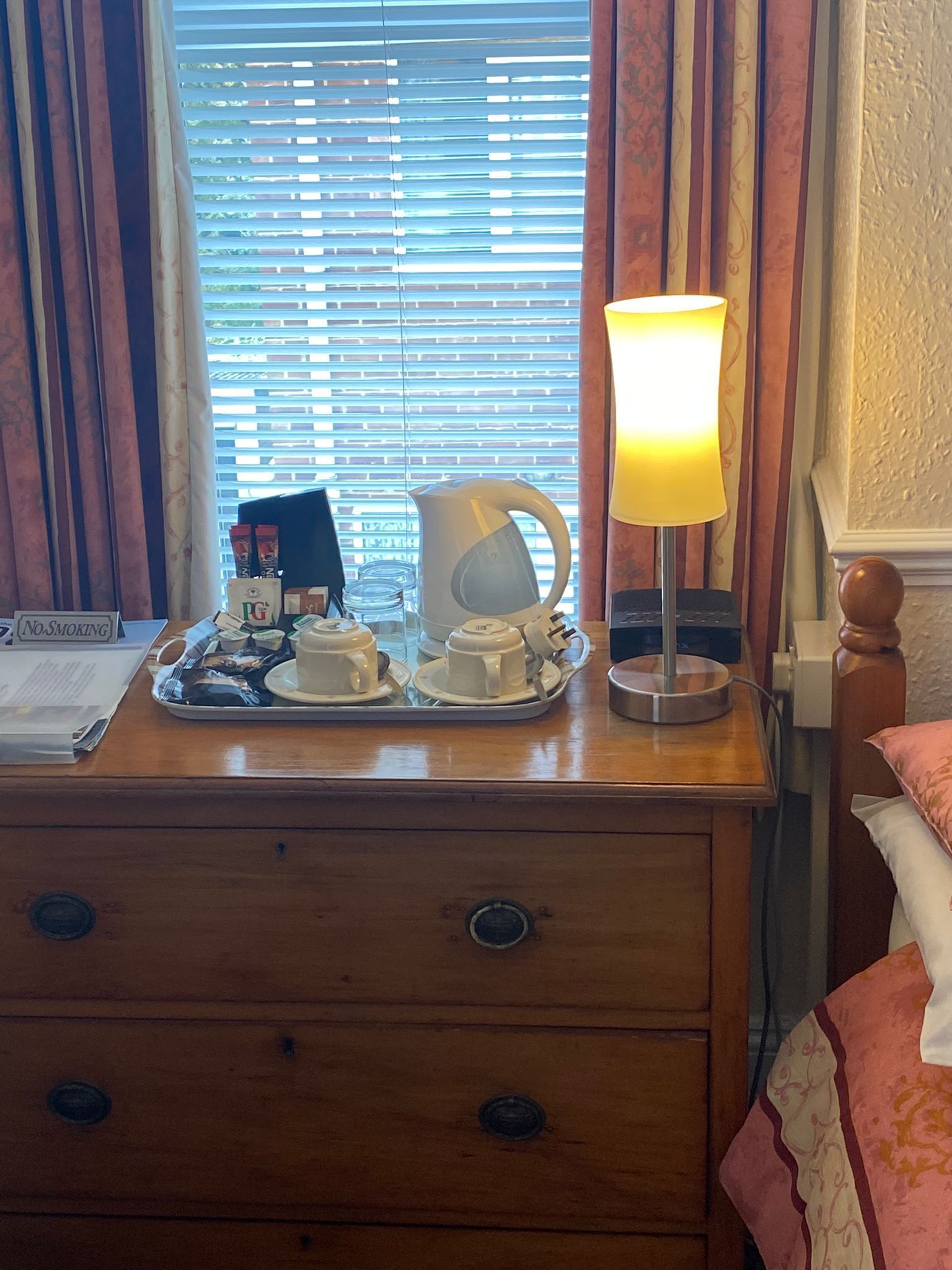 A wooden nightstand with a tray of white cups and saucers, a white electric kettle, and a table lamp with a yellow shade.