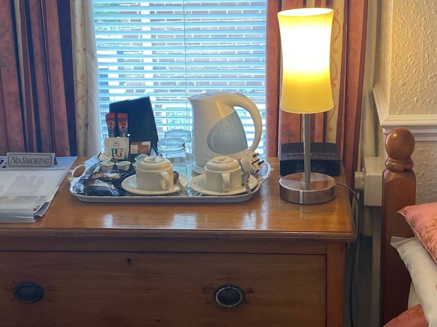 A wooden nightstand with a tray of white cups and saucers, a white electric kettle, and a table lamp with a yellow shade.