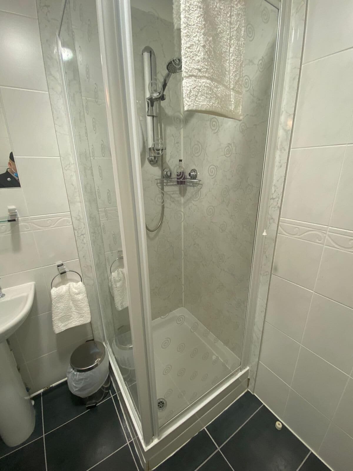 A glass-enclosed shower with a white towel hanging on the door, a sink, and a trash bin.