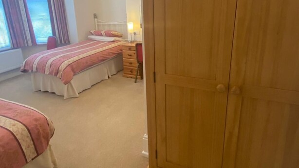 A bedroom with two beds, a wooden wardrobe, a bedside table with a lamp, and a window with curtains. - Landguard Lodge (Southampton)